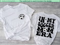 Two white t-shirts with soccer-themed designs on a wooden surface.